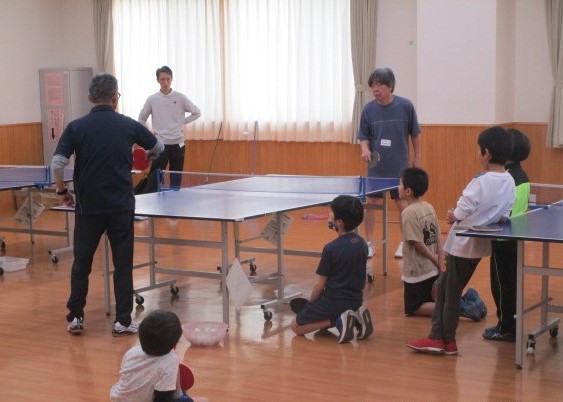 卓球教室で、指導者がラケットとボールを使って打ち方の実演をしており、子供たちが見守っている様子を写した、卓球指導の写真