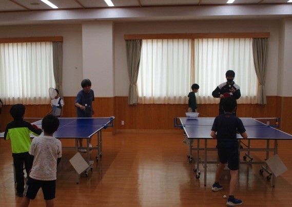 卓球教室で、複数の卓球台を使って子供たちが練習しており、指導者がそれぞれのグループを見て回っている様子を写した、卓球教室活動の写真