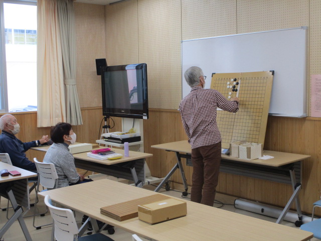 囲碁の盤を前にして講師が説明をしており、参加者が真剣に耳を傾けている様子を写した、地域囲碁教室の写真