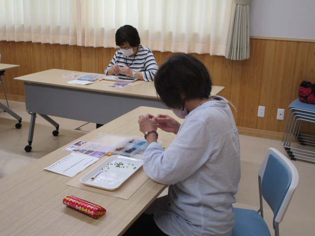 明るい室内で材料や道具が広げられた机上で、手前の女性と奥の女性も同様の作業をしている写真