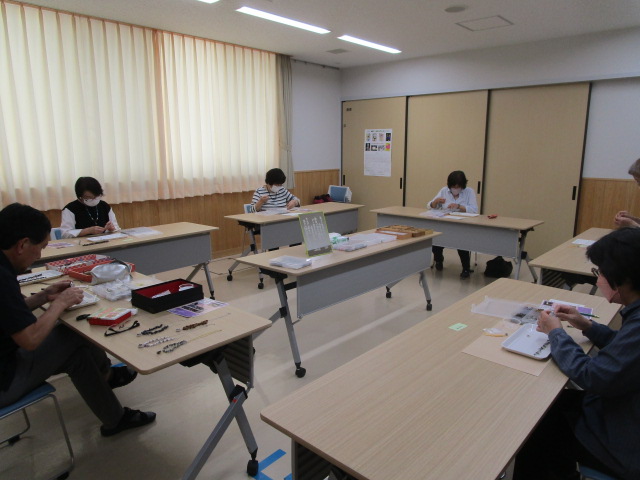 公民館のような室内で、数名の参加者がそれぞれ間隔を空けて配置された机に向かい、ビーズなどの手芸材料を広げて真剣に作業をしている様子の写真