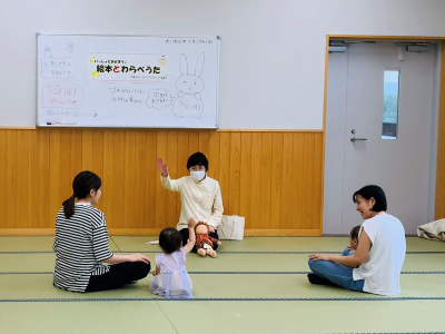 畳の部屋で、白いボードの前に座っている人が手を挙げて話しており、周囲には子供と一緒に座っている大人がいる様子を写した、地域交流イベントの写真
