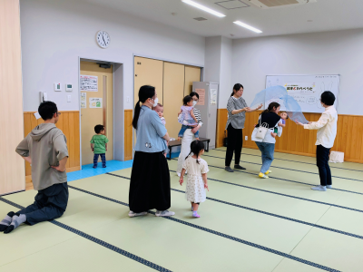 畳の部屋で、大人たちが子供を抱えたり見守ったりしながら活動しており、一部の人が青い布を持ち上げて遊んでいる様子を写した、親子ふれあい遊びの写真