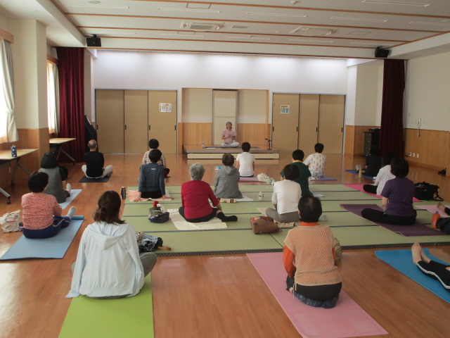 木造の床と畳が敷かれた広い和室で、中央の壇上で座禅を組む講師に向けて、床に敷かれたヨガマットや座布団に座り瞑想やヨガの指導を受けている様子の写真
