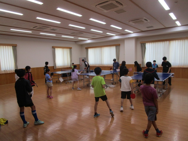 子供たちが卓球台を囲んでラケットを持ち、楽しそうに卓球をしている様子を写した、子供向け卓球教室の写真