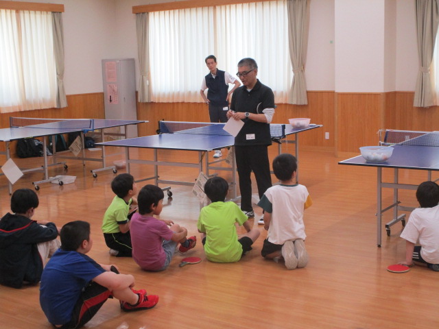 複数の子供が卓球台の周りに座り、大人が立って何かを説明している卓球教室の写真