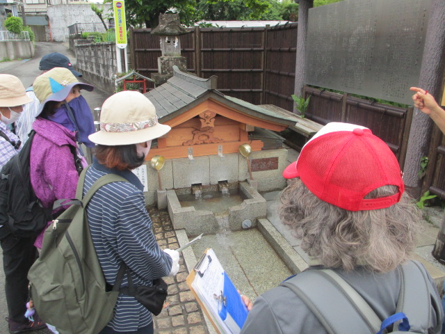 薄暗い曇り空の下、帽子をかぶった5人の人々が、木組みの屋根を持つ石造りの小さな湧き水、または手水舎のような施設を囲んでおり、一人がメモを取っている様子を捉えたスナップ写真