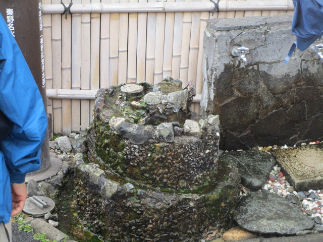 竹垣を背景に、苔むした三段の石組みから湧き水が流れ落ちている自然の湧水、右奥に蛇口が付いた大きな石がある光景を写した屋外写真