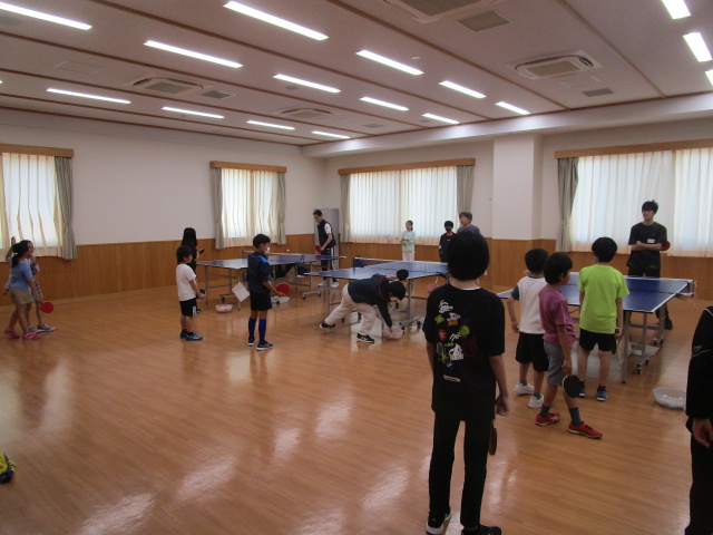 子供たちが卓球のラケットを持って打ち合いをしており、周囲には順番を待つ子供たちが並んでいる様子を写した、卓球教室の写真