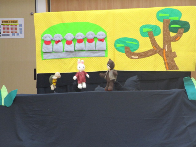 黄色い背景の舞台上で、ウサギとクマとカメの人形が並び、背景には緑の木と赤いスカーフを巻いた五体の地蔵らしき人形が並ぶ様子を描いた布製の飾り付けによる人形劇の舞台の写真
