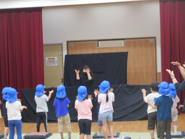 青い帽子をかぶった複数の園児たちが黒い幕の前で両手を上げて立ち、先生と思われる人が幕の後ろから身を乗り出し顔の前で両手を広げてジェスチャーをしている様子を後ろから捉えた写真