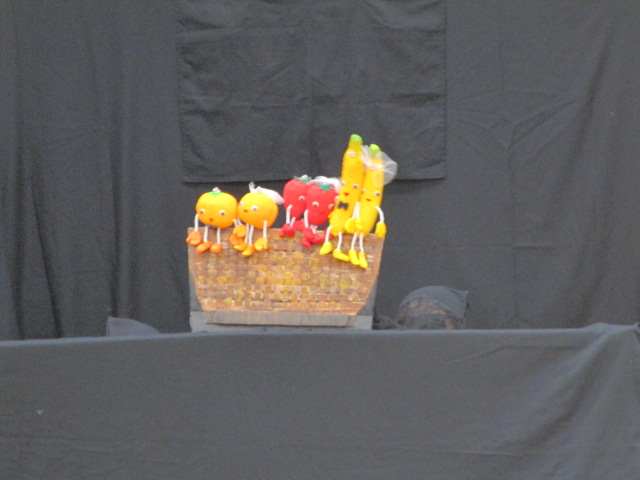 黒い幕が張られた舞台の上で、カゴの中にオレンジ色の顔に手足がついたミカン、赤い顔のトマト、黄色いバナナといった果物と野菜のキャラクター人形が並べられている様子の写真