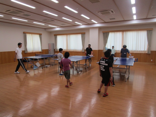 大きな窓と明るい照明の木目調の広間で、卓球台が複数設置され、数名の大人と子どもが卓球を楽しんでいる様子の写真