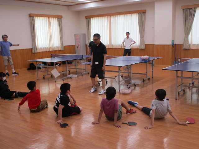 窓と照明が多数ある明るいフローリングの広間に、卓球台が3台設置され、子どもたちが床に座っている前で大人が何かを説明している写真