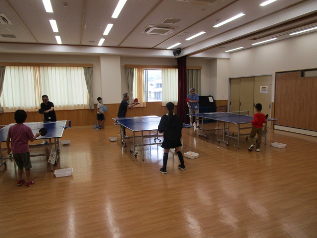 窓から自然光が差し込み、天井の蛍光灯が明るい広々としたフローリングの空間に、複数の卓球台が設置され、大人と子どもがそれぞれ熱心にプレイしている様子の写真
