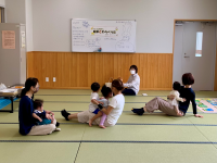 明るい和室のような空間で、数名の母親が幼い子どもたちを抱きかかえたり、近くで遊ばせたりしながら和やかな雰囲気で交流している様子の写真