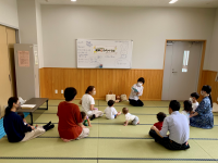 白い壁と木目調の腰壁の和室のような空間で、数名の大人たちが床に座り、数人の乳幼児を交えて和やかな雰囲気の中で交流する様子を捉えた写真