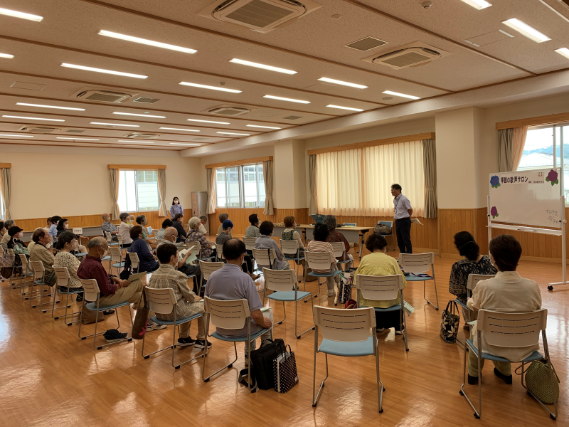 照明の整った広いフローリングの部屋で、大勢の高齢者が椅子に座って前方を見つめ、右奥で男性が立って説明している講演会の様子の写真