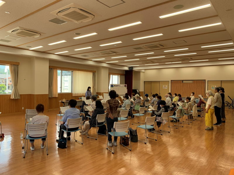 明るいフローリングの広間全体に、多数の高齢者が椅子に座り楽譜のような紙を持って合唱練習に参加している集合写真