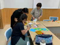 白い壁と木目調の腰壁の部屋で、3人の女性がテーブルを囲み、ハサミや色紙などを広げて七夕かざり作りをしている作業風景を捉えた写真