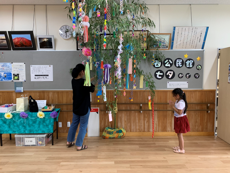 室内で、女性が緑の笹に吊り下げられた色とりどりの七夕飾りを調整し、少女が手を合わせて願い事をする様子を捉えた写真