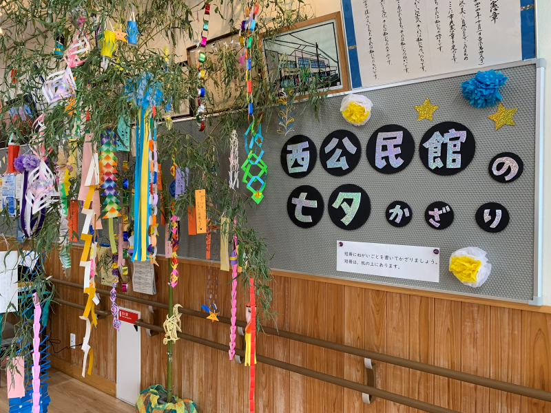 緑の笹に色とりどりの短冊や吹き流しなどの七夕飾りが吊るされ、その奥の壁には「西公民館の七夕かざり」と書かれた掲示板がある館内の写真