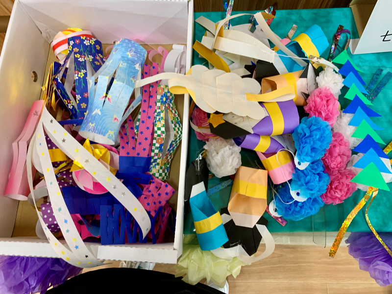 白色の箱の中に星柄や和柄の折り紙で作られた輪飾りやちょうちんなどの七夕飾りと、テーブルの上に青やピンクのボンボンや黄色い短冊状の飾りが山積みになった作業中の写真