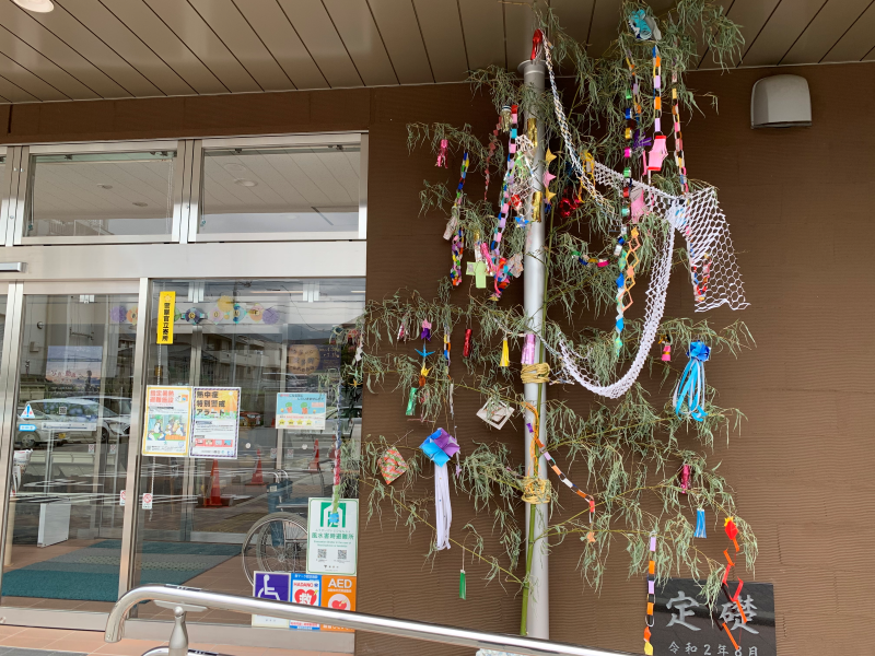 ブラウンの壁の建物入り口前に、細長い竹にカラフルな短冊や七夕飾りが取り付けられている写真