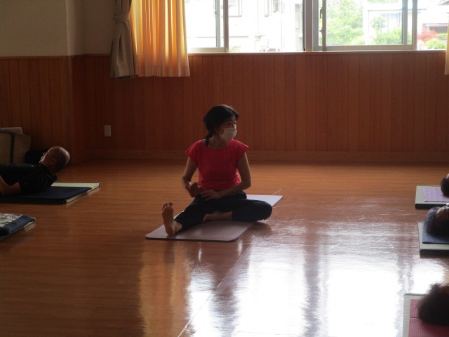 フローリングの広間で、一人の女性がマスクを着用し、赤いTシャツと黒いズボン姿でヨガマットの上に座り、片足を伸ばすポーズをしている運動中の写真