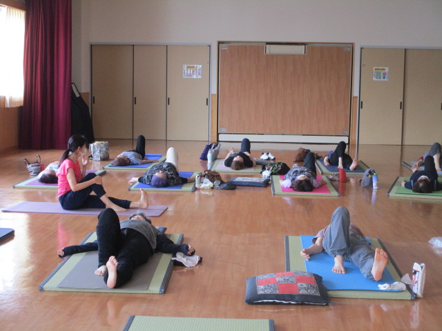 広いフローリングの部屋で、インストラクターの指導のもと、参加者たちが各自のヨガマットや畳の上で仰向けになるリラックスしたポーズをとっている運動中の写真