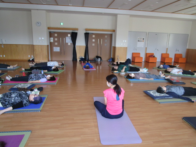 明るいフローリングの広間全体に多くの参加者がヨガマットや畳の上で仰向けになり休息する中、ピンクのTシャツを着た指導者が座っている運動中の写真