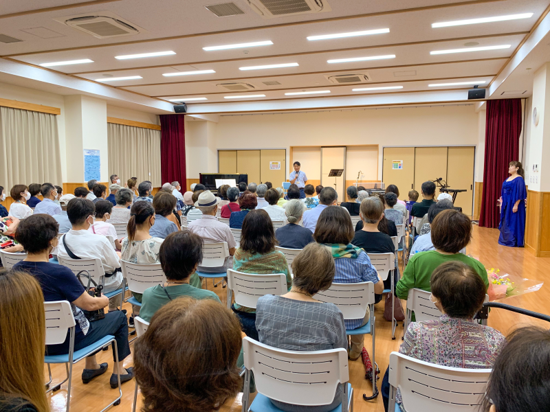 多数の聴衆が白い椅子に座って前方を見つめ、舞台上では男性がマイクを持って話をしており、舞台袖に青いドレスの女性が立っている写真