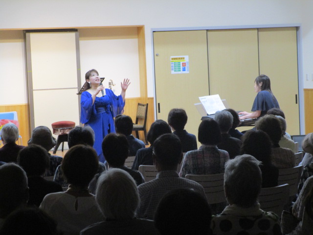 青いロングドレスを着用した女性がマイクを持って歌い、右奥で別の女性がピアノを演奏しており、多くの聴衆が後ろ姿で鑑賞している室内コンサートの様子の写真