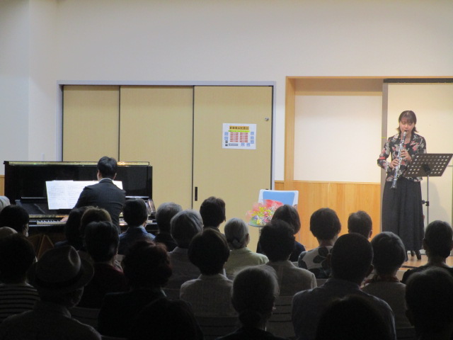 舞台の上で女性がクラリネットのような木管楽器を演奏しており、左奥では男性がピアノを弾き、多数の聴衆が鑑賞している室内での音楽会の写真