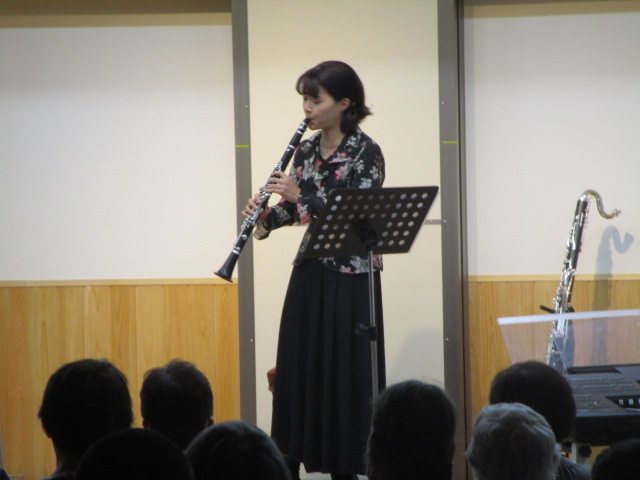 黒いワイドパンツに花柄のブラウスを着用した女性が、ステージ上で譜面台を前に、クラリネットを演奏している写真