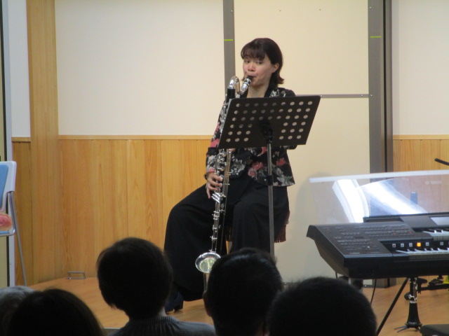黒いワイドパンツと花柄のブラウスを着用した女性が、譜面台の前に座り、大型のバスクラリネットを演奏している音楽会の写真