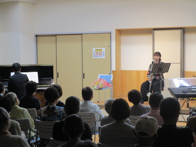 和室の襖のような引き戸と白い壁の部屋で、女性の大型のバスクラリネットの演奏と男性のピアノの演奏を多数の聴衆が鑑賞しているコンサートの様子の写真