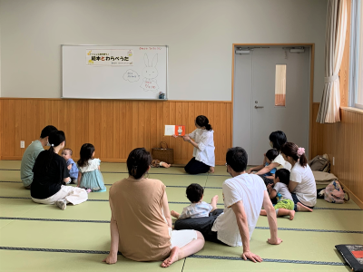 畳の部屋で数組の親子が輪になって床に座り、中央で女性が絵本を広げて絵本の読み聞かせをしている様子を捉えた写真