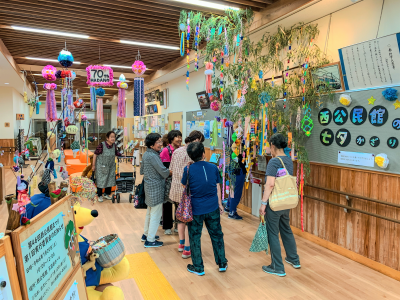 広々とした木目調の館内に、カラフルな七夕飾りが多数吊るされ、その一角に集まり談笑する数名の高齢者たちがいる賑やかな様子の写真