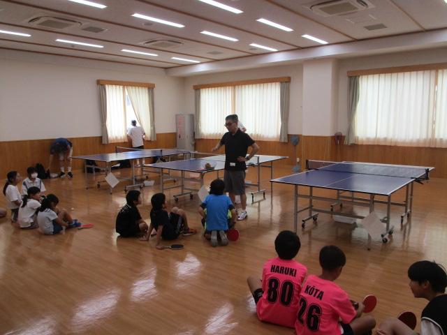 光沢のあるフローリングの室内で、数台の卓球台を設置し、大人の指導者が床に座る多くの子どもたちに何かを説明している卓球教室の写真