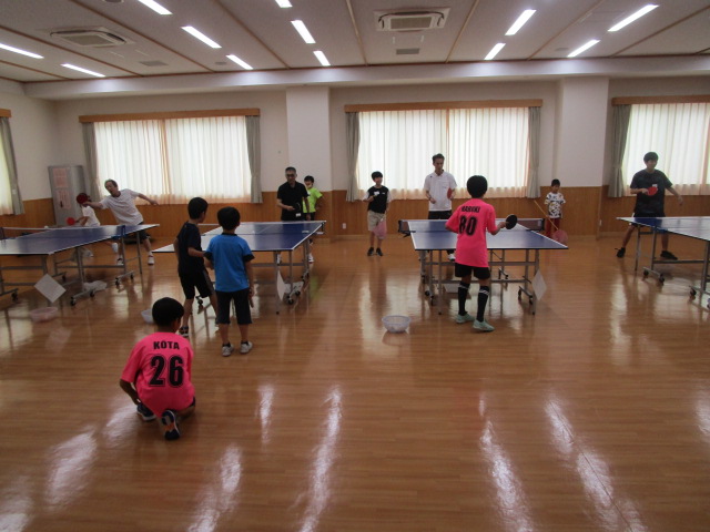 光沢のあるフローリングの室内で、数台の卓球台を設置し、大人の指導者が見守る中子供たちがラリーを続けている卓球教室の写真