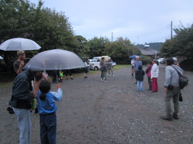 砂利の駐車場で、数人の大人と子どもが傘をさしたり、ビニール傘を頭上に掲げたりしながら集まっている写真