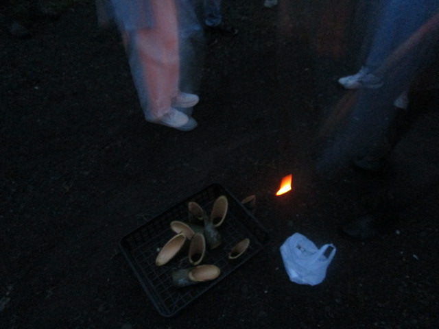 薄暗い屋外で、竹を加工して作られた器が金属製の網の上に置かれており、その横の地面には小さな炎が上がっている夜間のイベント風景をクローズアップした写真