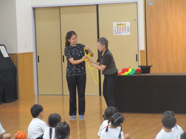 フローリングの室内で、2人の女性が黄色い縄を輪にして持つマジックを披露し、多くの幼児が床に座ってそれを熱心に見つめているレクリエーション風景の写真