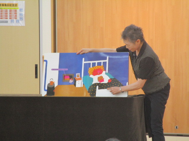 女性が黒い布で覆われた台の前で、読み聞かせ用の絵本のようなボードを指さして話をしている写真