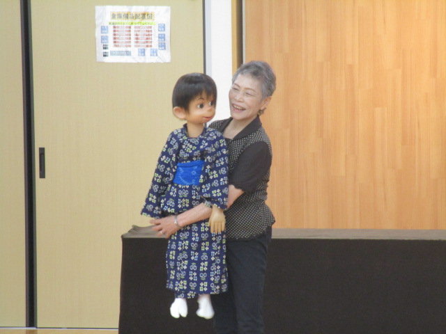 和服姿の少年型人形を抱きかかえた女性が、笑顔で前方を向いている腹話術の様子の写真