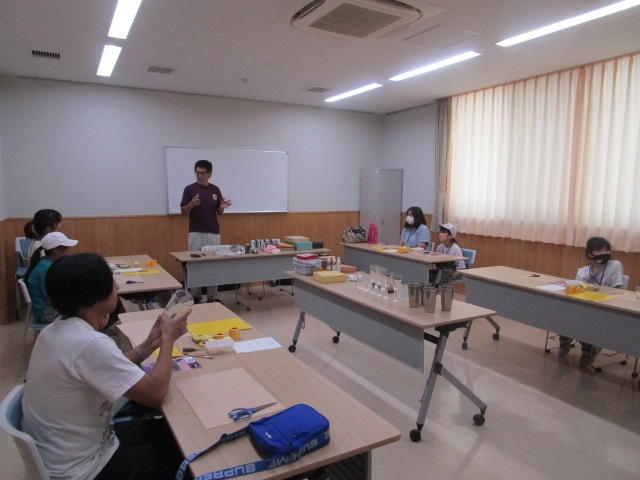 白板の前で講師の男性が参加者に向けて話しており、各テーブルにはグラスやハサミなどが置かれ、数名の子どもと大人が着席しているワークショップ風景の写真