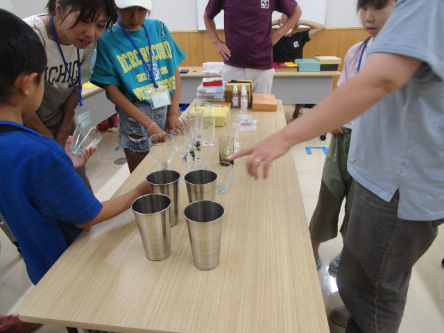 作業机を囲んだ数人の子どもたちと大人が、テーブルの上に並べられたガラスのコップと金属製のタンブラーを指さしながら何かを話し合っているワークショップ風景を近接撮影した写真