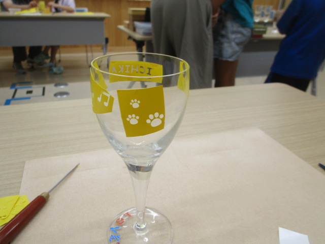 机の上に置かれたワイングラスのような脚付きの透明なグラスに、黄色いシールで「ICHIKA」の文字と音符、そして犬の肉球の模様が貼り付けられている制作途中の作品の写真