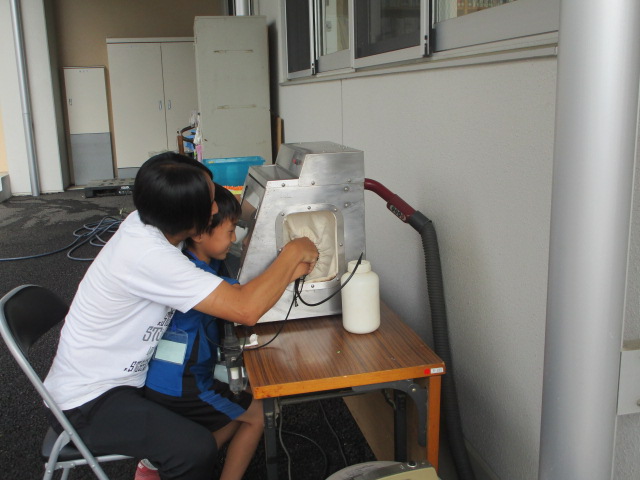 屋外の軒下のような場所で、白いTシャツを着た大人が、男の子を膝に乗せ、金属製の箱型の機械に手を入れて中の作業を見守りながら作業している様子の写真